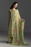A woman models a traditional three-piece salwar kameez suit in sage green with white floral embroidery, paired with a matching sheer dupatta featuring pink and green abstract patterns, set against a dark gray studio background.