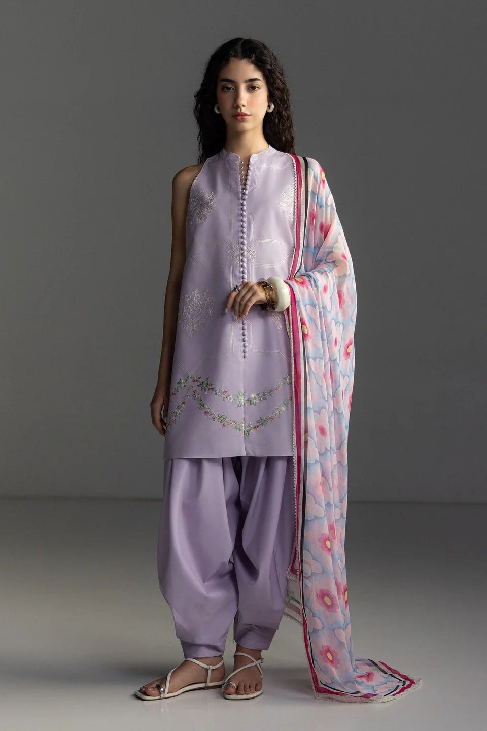 Woman wearing a traditional outfit with a light purple kurta, pants, and a floral dupatta against a gray background.
