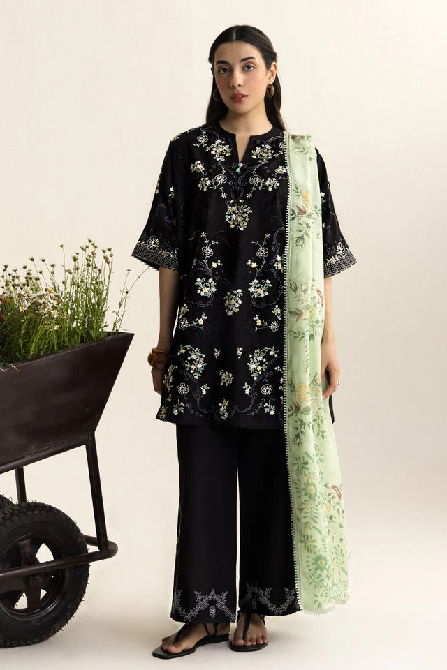 Woman wearing a black floral outfit with a green scarf, standing next to a wheelbarrow with plants.