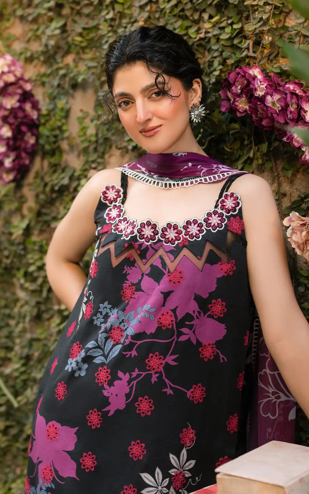 A woman wearing a sleeveless black dress with vibrant pink, purple, and red floral embroidery, paired with a matching purple scarf, posed against a background of green ivy and purple flowers.