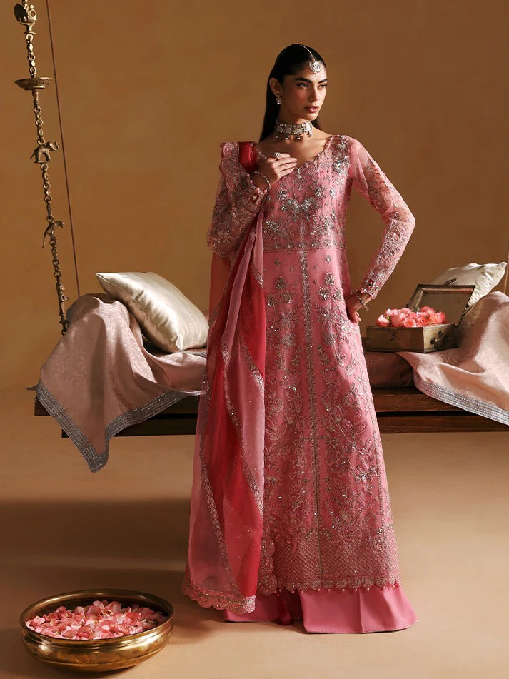 Woman in a pink embroidered traditional outfit standing in a decorated room with a swing and pillows.