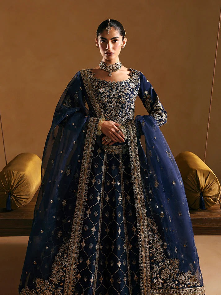 Woman wearing a traditional blue embroidered outfit with gold details against a beige background.