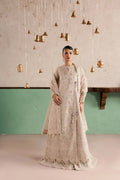 A woman stands in a studio wearing a long beige embroidered traditional outfit with a matching draped shawl. The background features a textured light wall with hanging brass bell decorations and a mint green lower panel.