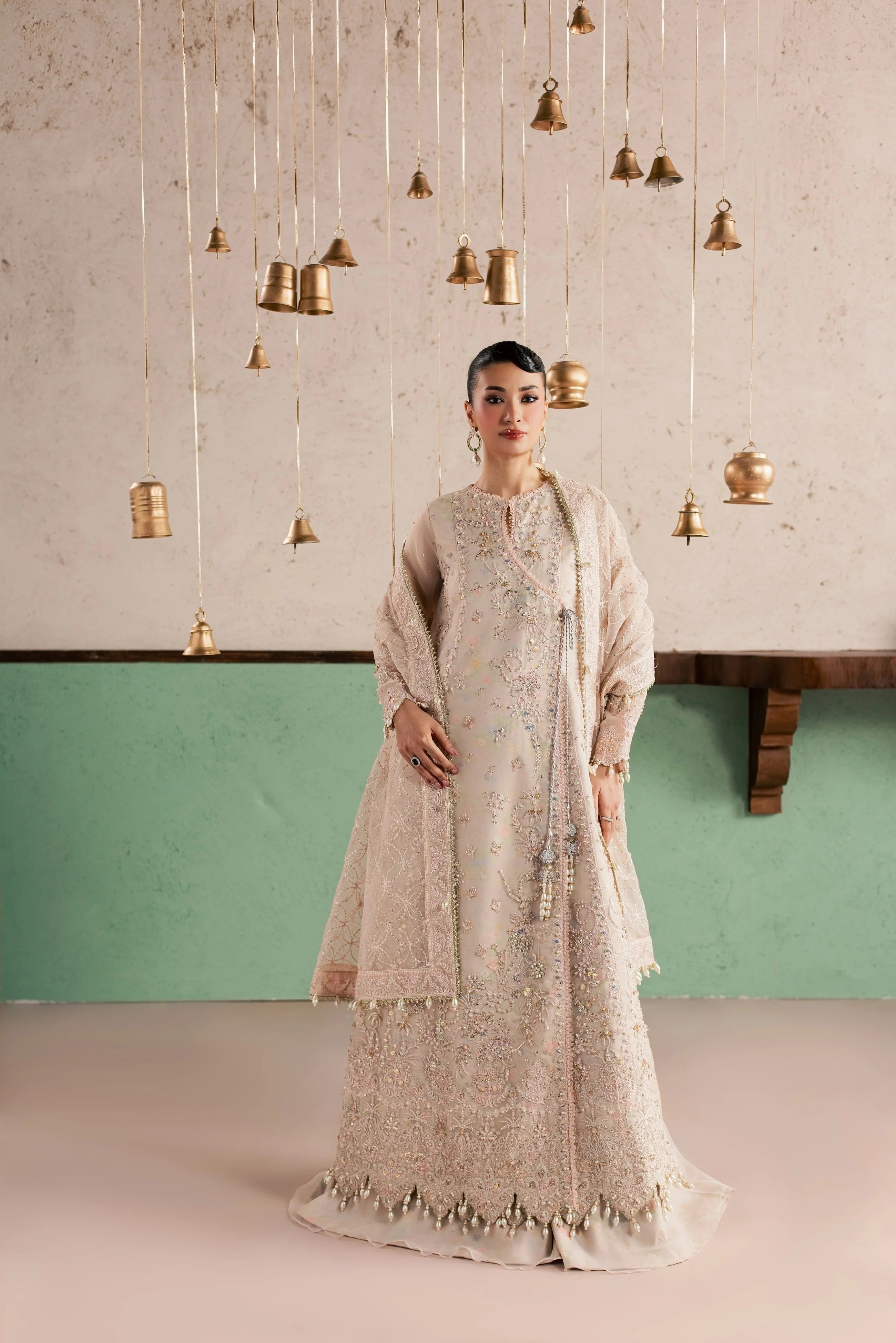 A woman stands in a studio wearing a long beige embroidered traditional outfit with a matching draped shawl. The background features a textured light wall with hanging brass bell decorations and a mint green lower panel.