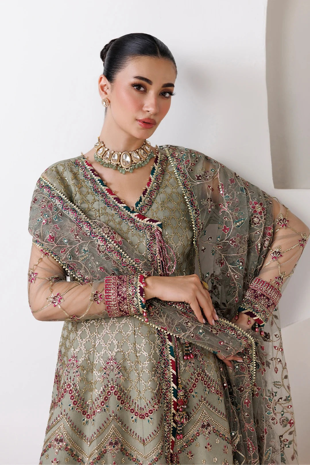 A woman models a traditional South Asian outfit featuring a heavily embroidered olive green kurta with pink and gold floral patterns, a matching sheer dupatta, and a chunky gold choker necklace.