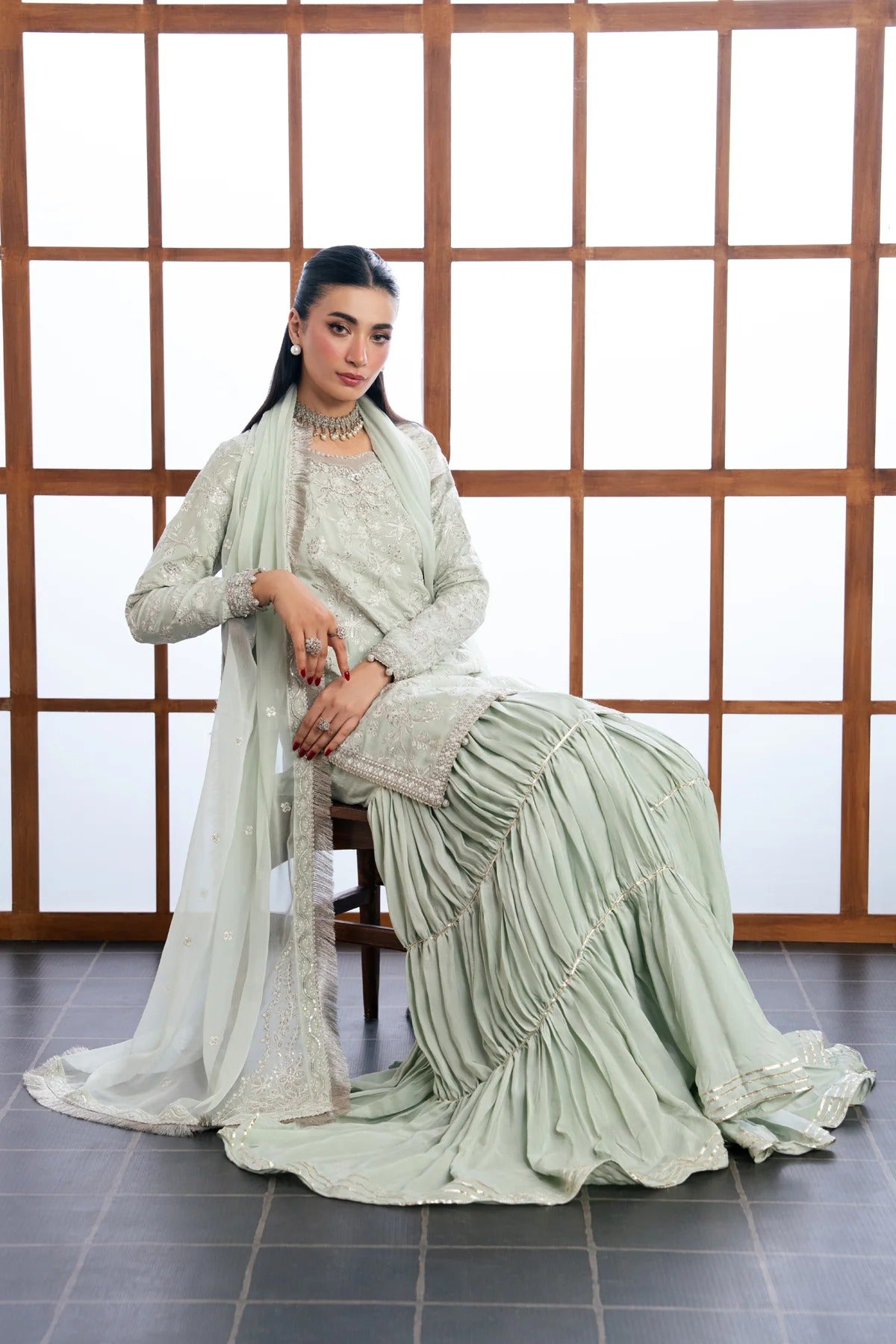 A woman models a light sage green traditional outfit with matching dupatta, seated on a wooden chair in front of a wooden grid window.