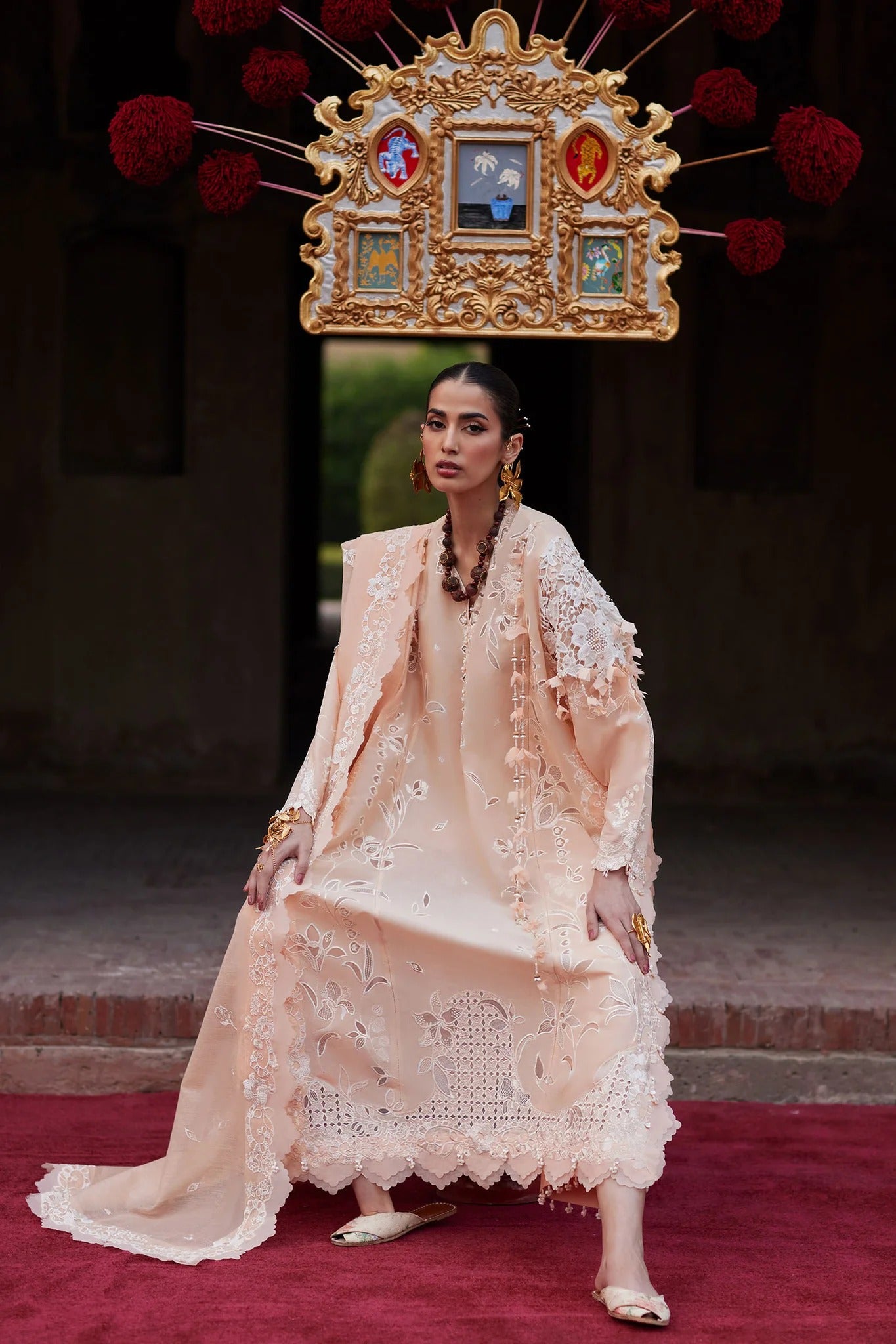 Woman in a beige embroidered outfit standing on a red carpet with decorative background