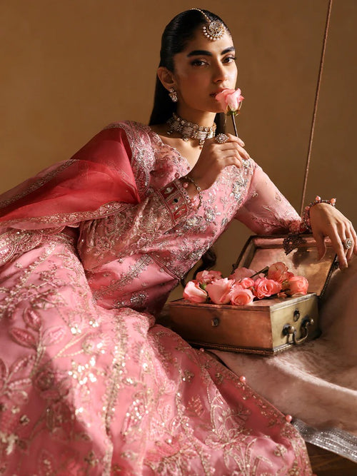 Woman in a pink traditional outfit with floral decorations, sitting on a beige background.