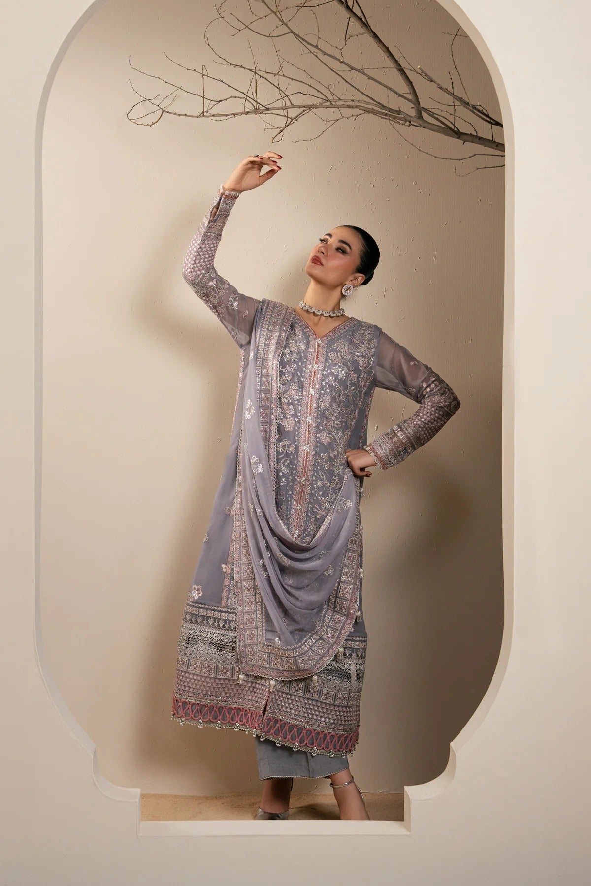 A woman models a traditional South Asian salwar kameez suit in a dusty lavender-gray color with intricate silver and gold embroidery, paired with a matching dupatta, set against a beige arched wall with decorative branches.