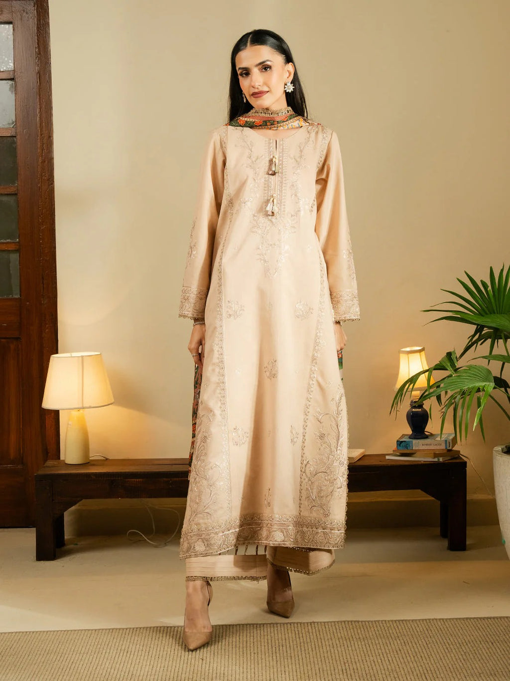 A woman modeling a long beige embroidered kurta with matching trousers and a dupatta in a warm, decorated indoor setting.