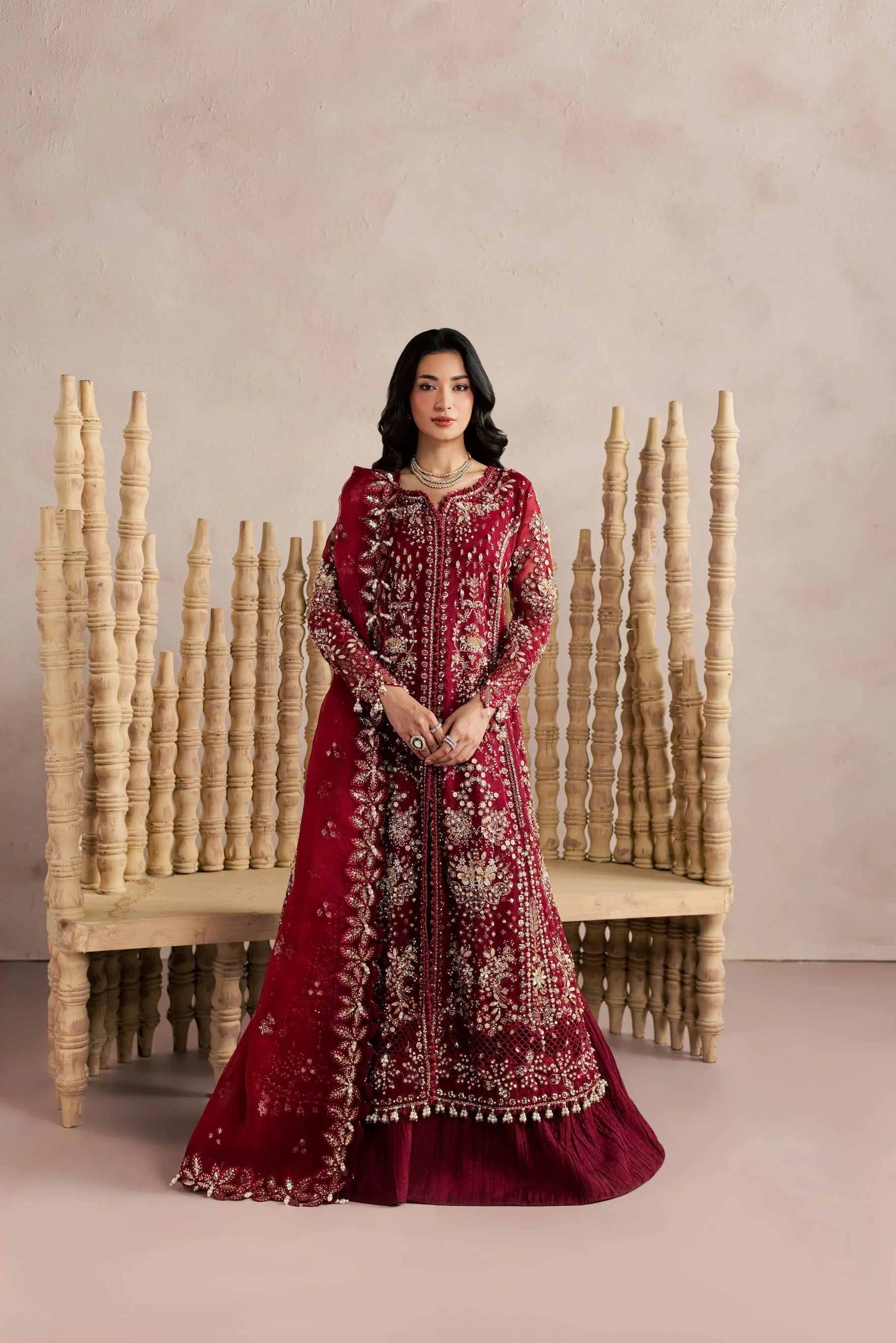 A woman stands in front of a beige wall and a carved wooden bench, wearing an ornate deep red traditional outfit with extensive gold embroidery and a matching embroidered dupatta.