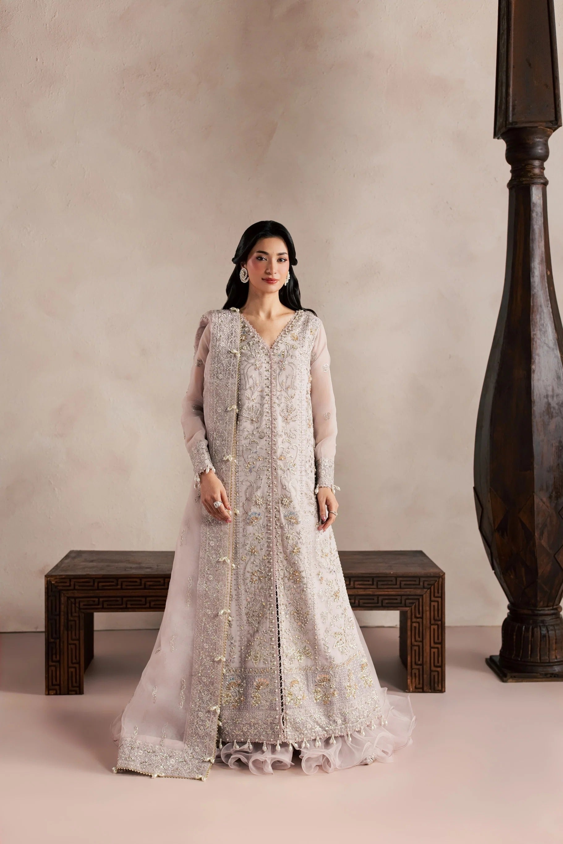 A woman stands indoors wearing a long, heavily embroidered light gray or silver traditional outfit with sheer sleeves and a sheer draped overlay. She is posed in front of a beige wall with a dark wooden bench and a large dark wooden column.