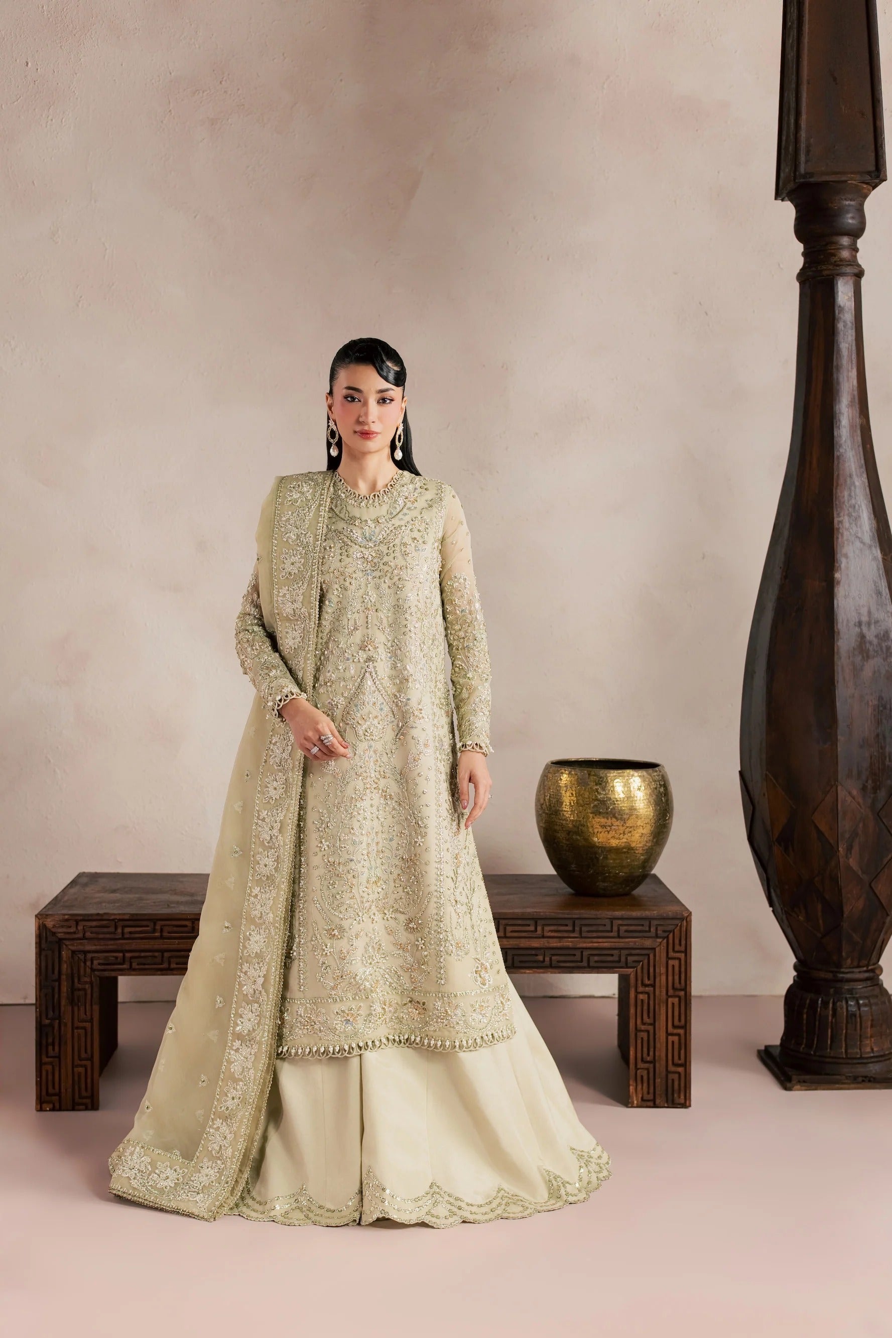 A woman models a traditional South Asian outfit consisting of a heavily embroidered light beige kurta, a matching dupatta, and beige palazzo pants. She is posed in a studio setting with a textured beige wall, a dark wooden bench, a gold decorative pot, and a large dark wooden pillar.