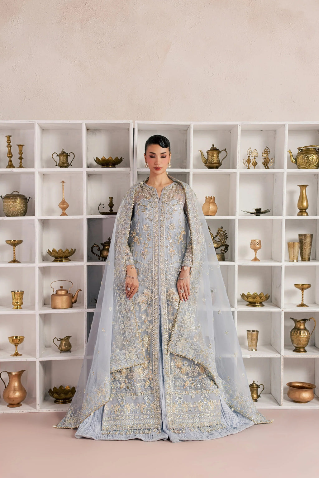 A woman stands in front of a white shelving unit filled with decorative brass and metallic objects, wearing a light blue heavily embroidered traditional outfit with a sheer draped overlay.