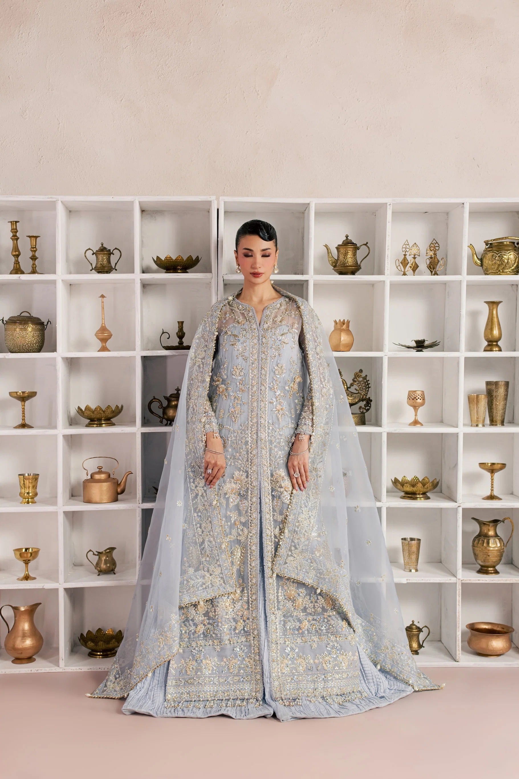 A woman stands in front of a white shelving unit filled with decorative brass and metallic objects, wearing a light blue heavily embroidered traditional outfit with a sheer draped overlay.