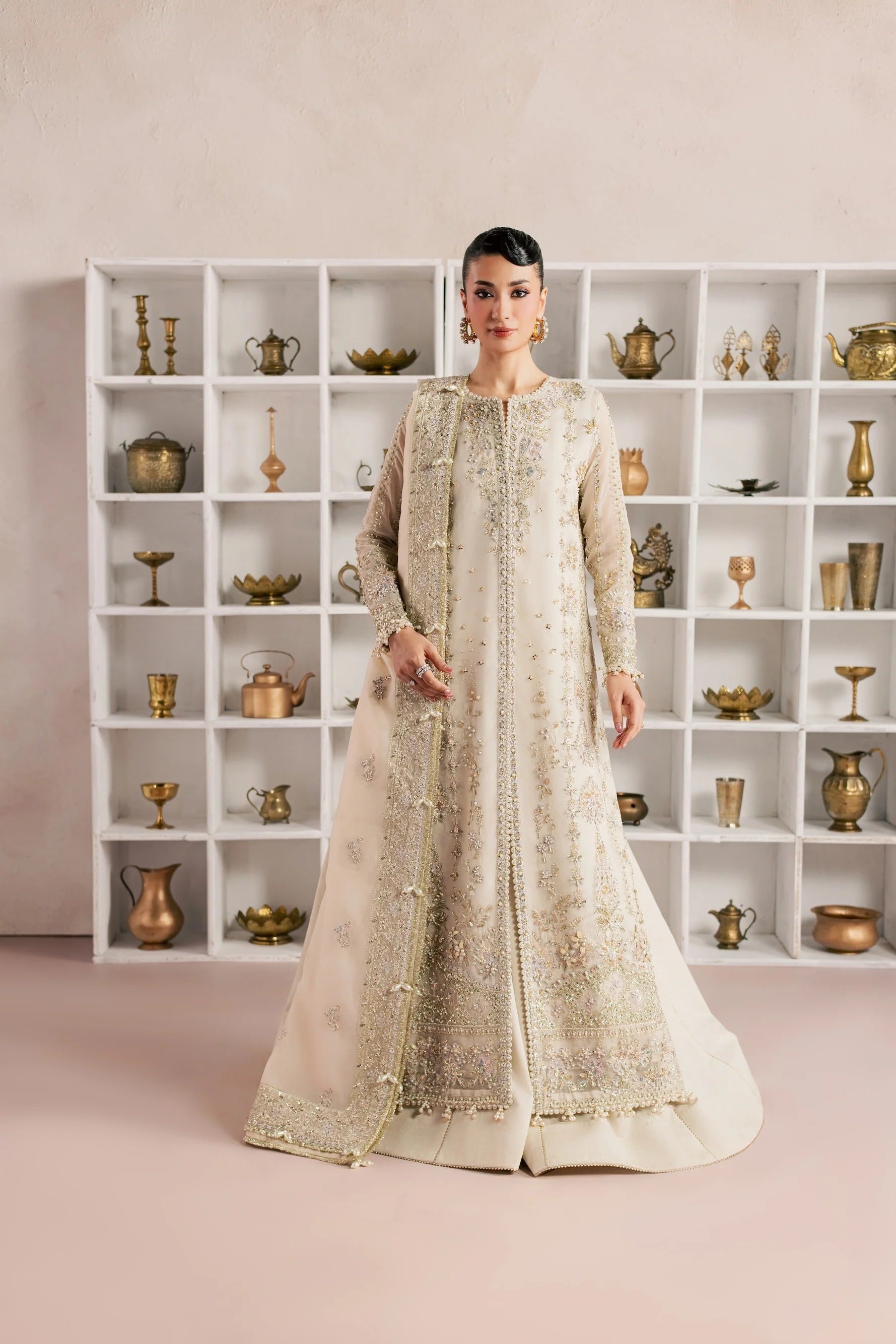 A woman stands in a studio wearing a long cream/off-white embellished traditional gown with gold embroidery and beadwork. She is posed in front of a white shelving unit filled with decorative brass and metallic objects against a soft beige-pink backdrop.