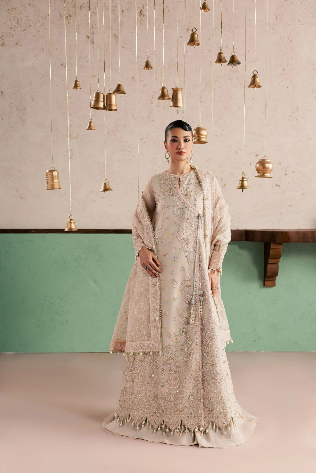 A woman stands in a studio wearing a long beige embroidered traditional outfit with a matching draped shawl. The background features a textured light wall with hanging brass bell decorations and a mint green lower panel.