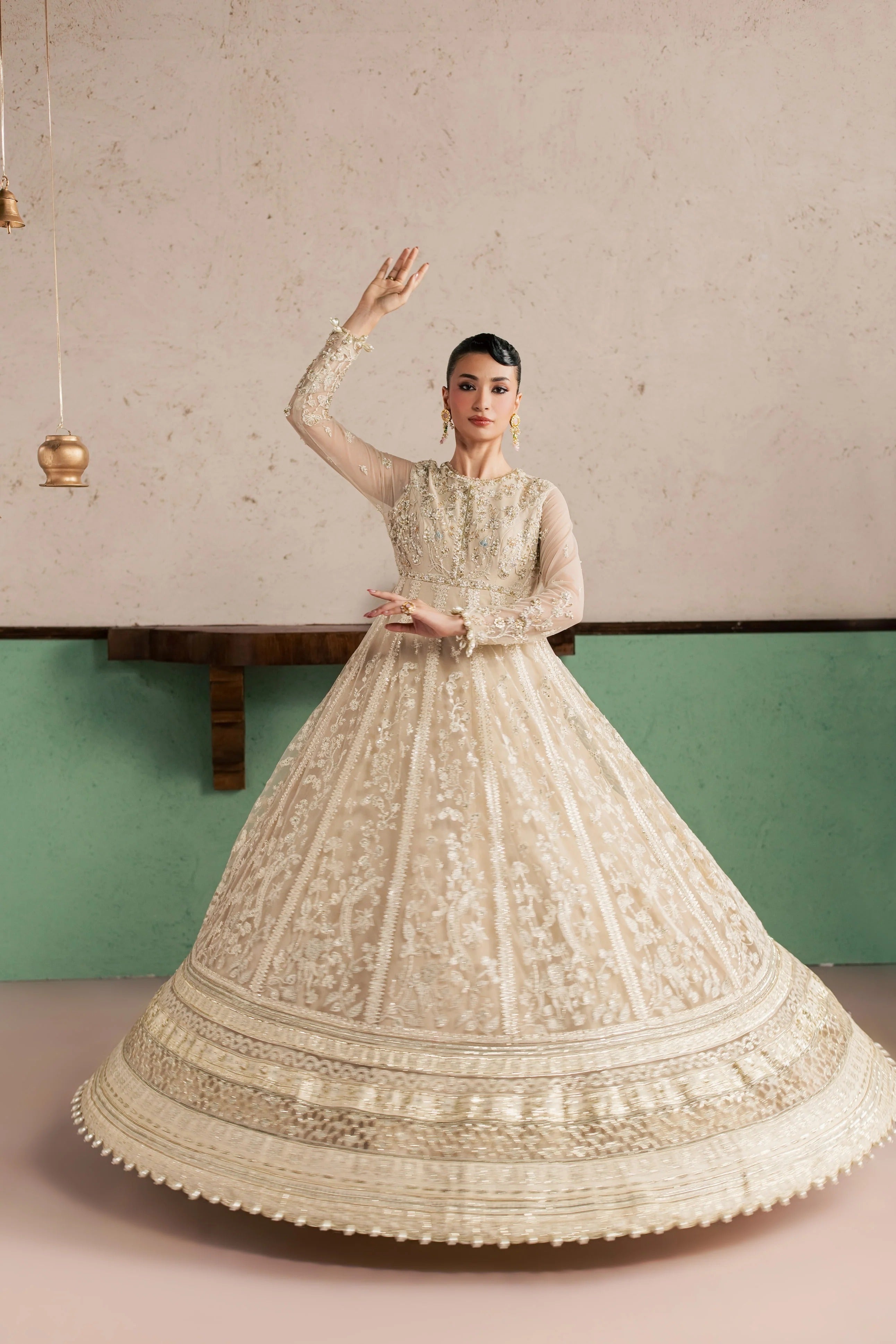 A woman modeling a voluminous cream-colored embellished gown with long sheer sleeves and intricate embroidery, posed against a textured beige wall and green lower wall panel.