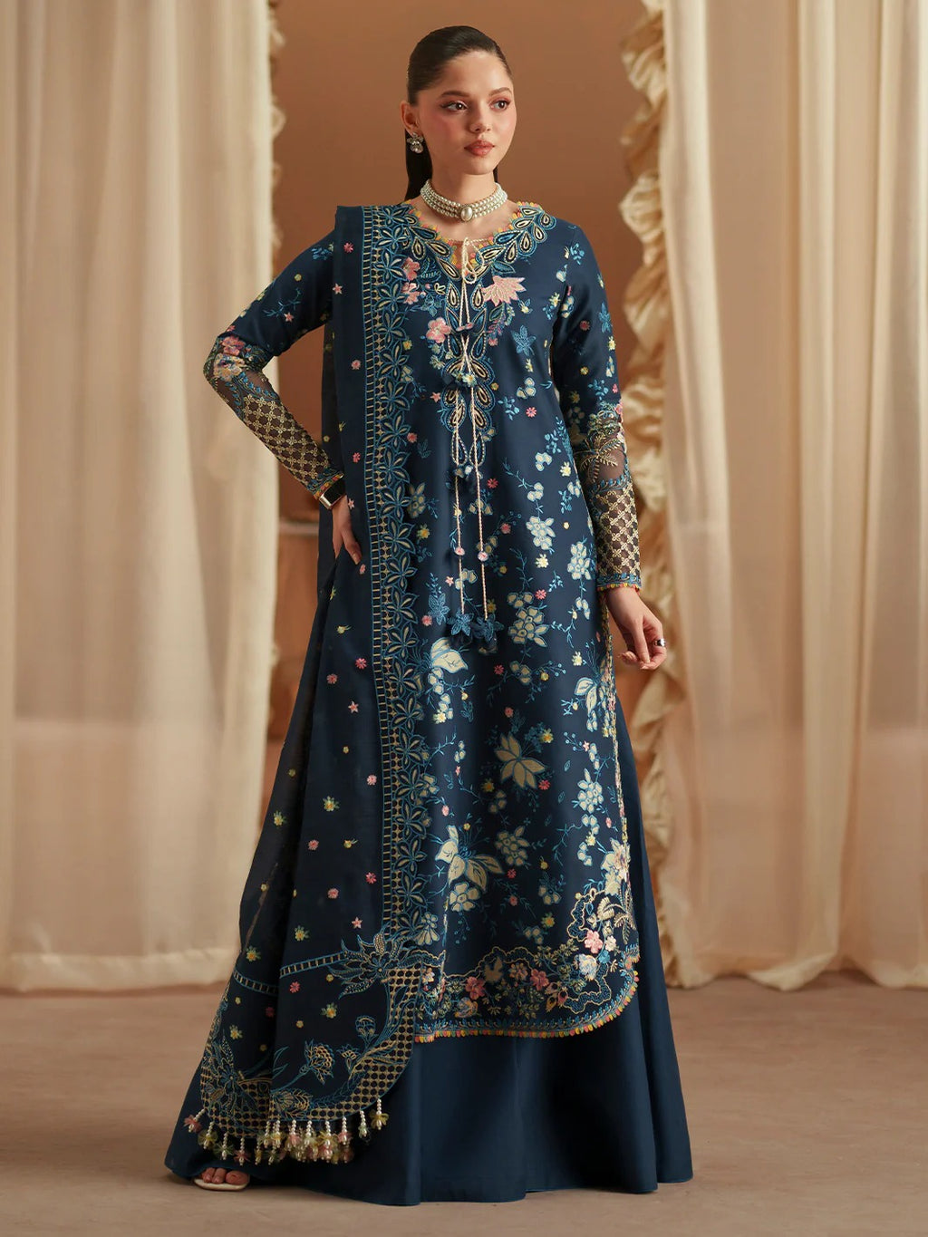Woman wearing a dark blue embroidered traditional outfit with gold details against a beige curtain background.