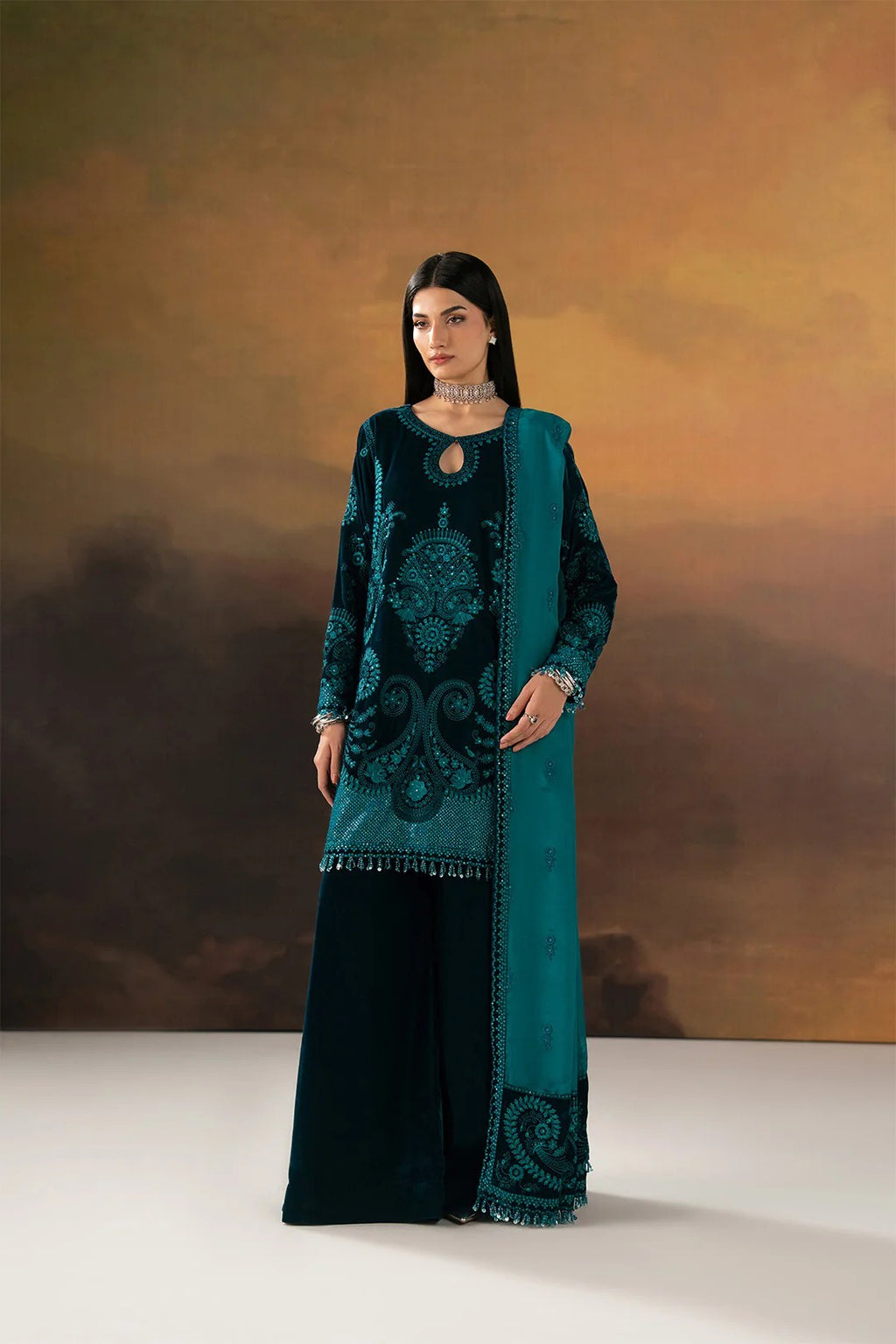 A woman modeling a traditional South Asian outfit consisting of a dark teal embroidered kurta, matching dupatta, and black wide-leg trousers, set against a warm brown and gold gradient studio backdrop.