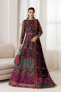 A woman models a traditional South Asian anarkali-style dress in deep plum purple with intricate gold embroidery, teal accents, and a matching embroidered dupatta, posed in a bright, modern living room setting.