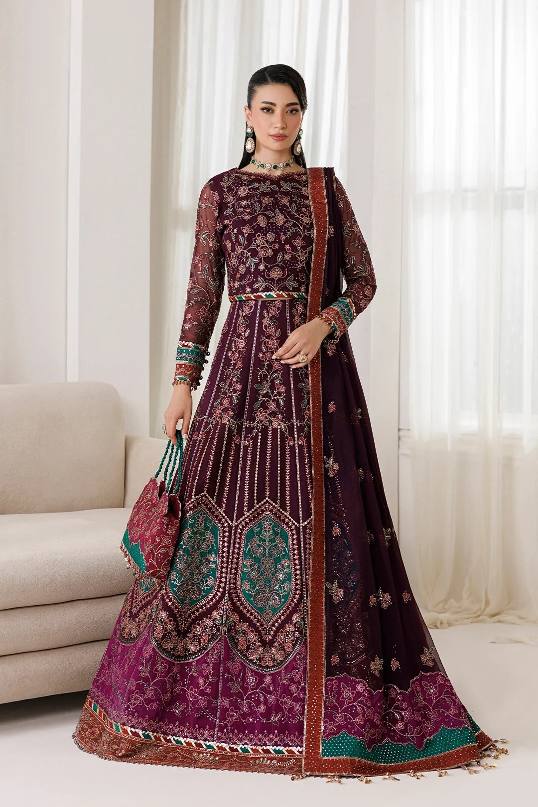 A woman models a traditional South Asian anarkali-style dress in deep plum purple with intricate gold embroidery, teal accents, and a matching embroidered dupatta, posed in a bright, modern living room setting.