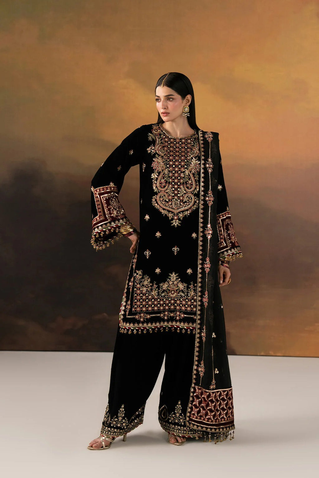 A woman models a black traditional outfit with extensive gold and red embroidery, featuring a long kurta, wide-leg pants, and a matching dupatta, set against a warm brown and gold studio backdrop.
