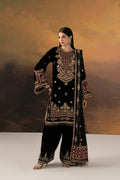A woman models a black traditional outfit with extensive gold and red embroidery, featuring a long kurta, wide-leg pants, and a matching dupatta, set against a warm brown and gold studio backdrop.