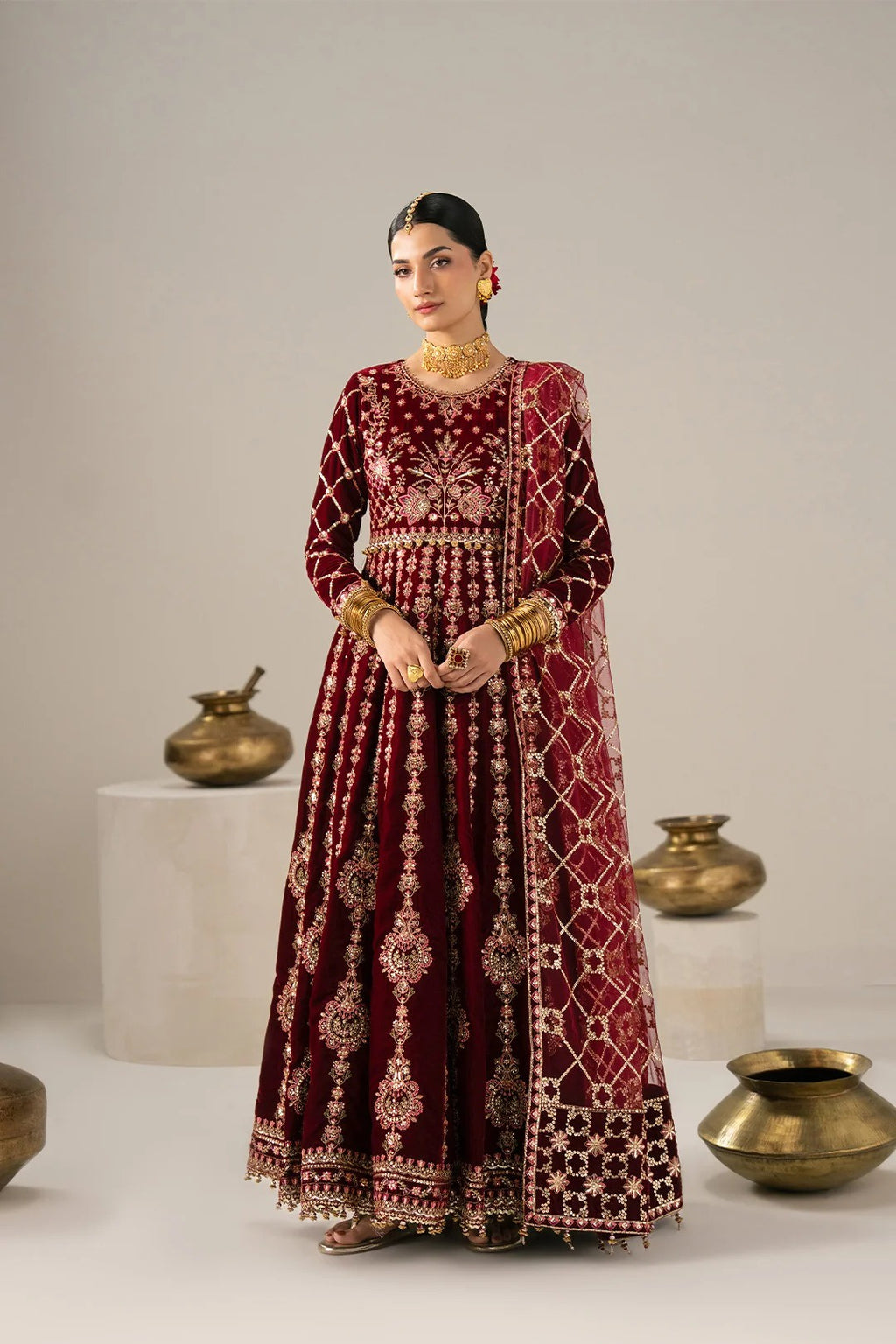 A woman stands in a studio wearing a long maroon velvet traditional outfit with extensive gold embroidery and a matching embroidered shawl. The setting has a neutral light gray background with decorative brass pots on white pedestals.
