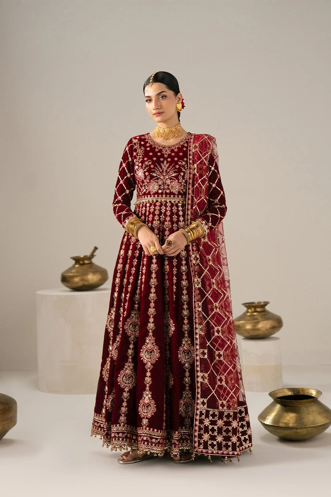 A woman stands in a studio wearing a long maroon velvet traditional outfit with extensive gold embroidery and a matching embroidered shawl. The setting has a neutral light gray background with decorative brass pots on white pedestals.