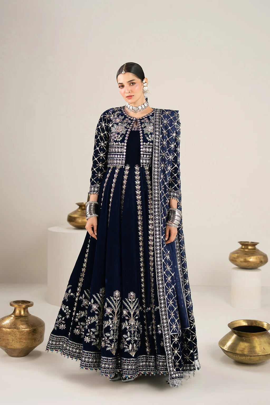 A woman models a dark navy blue traditional Anarkali gown with intricate silver embroidery and a matching embroidered jacket. The set is photographed in a studio with decorative brass pots on white pedestals against a light beige background.