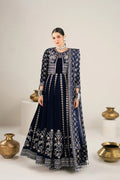 A woman models a dark navy blue traditional Anarkali gown with intricate silver embroidery and a matching embroidered jacket. The set is photographed in a studio with decorative brass pots on white pedestals against a light beige background.