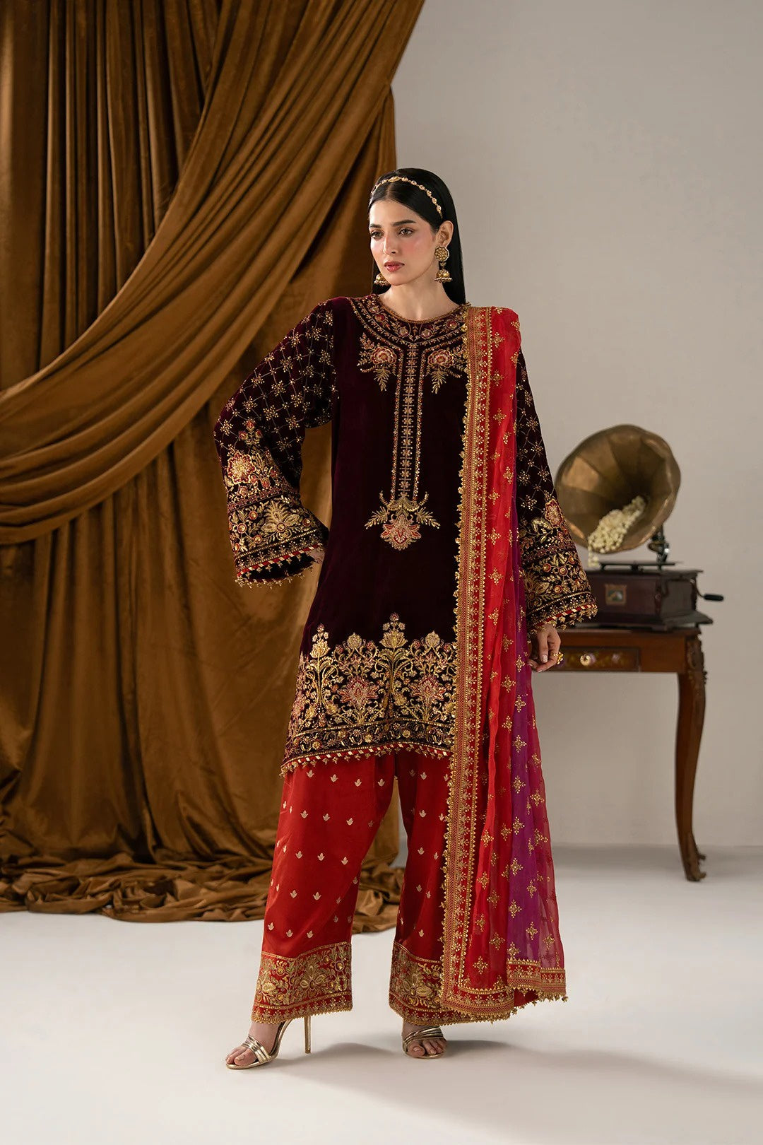 A woman models a traditional South Asian salwar kameez suit in a studio setting. The outfit features a deep maroon velvet tunic with intricate gold embroidery, paired with red wide-leg trousers and a red and purple dupatta decorated with gold patterns.