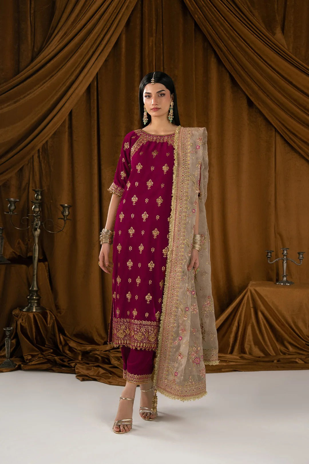 A woman models a traditional Indian salwar kameez suit in deep magenta with gold embroidery, paired with a beige printed dupatta, set against a draped brown velvet backdrop.