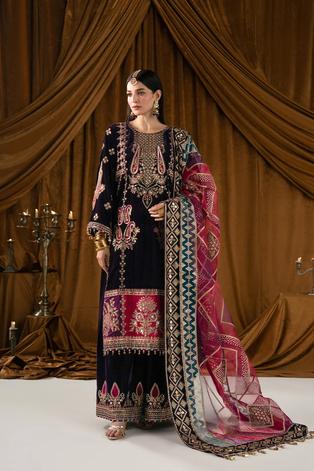 A woman models a traditional South Asian salwar kameez suit in dark navy blue with intricate gold, pink, and red embroidery, paired with a matching patterned dupatta, set against a draped brown curtain background.