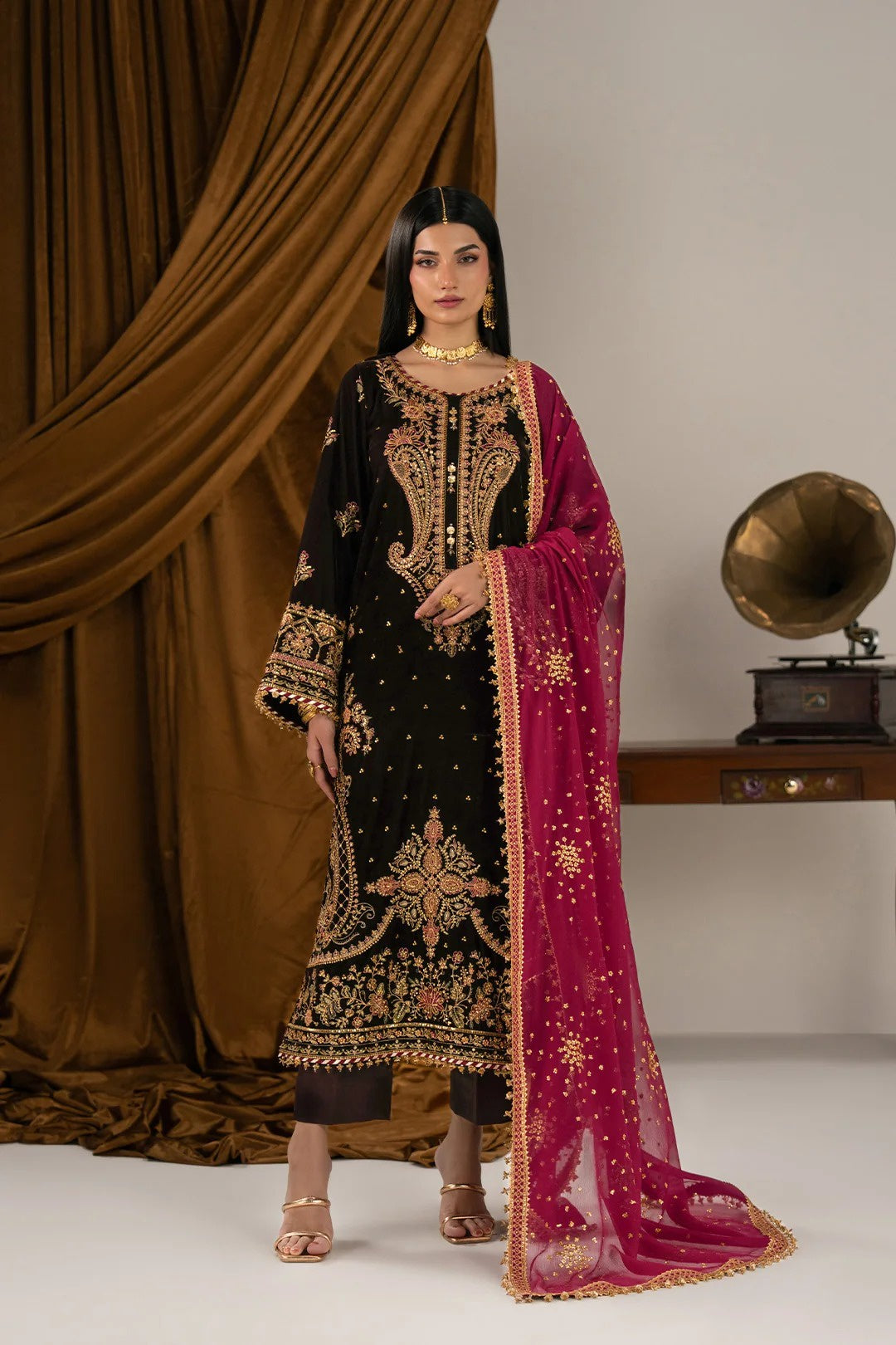 A woman models a traditional black and gold embroidered kurta set with a deep magenta dupatta draped over her shoulder, set against a backdrop of brown draped curtains and a vintage gramophone.