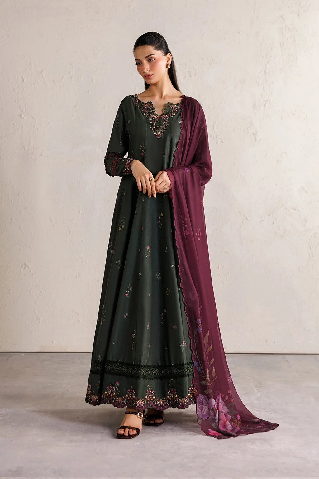A woman models a dark green embroidered Anarkali gown with a deep purple dupatta draped over one shoulder, set against a neutral textured wall.