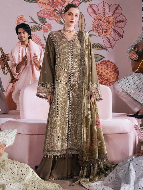 A model stands in a pink decorated room wearing an ornate olive green embroidered traditional outfit with a matching dupatta. Other people in light-colored clothing are partially visible in the background and foreground.