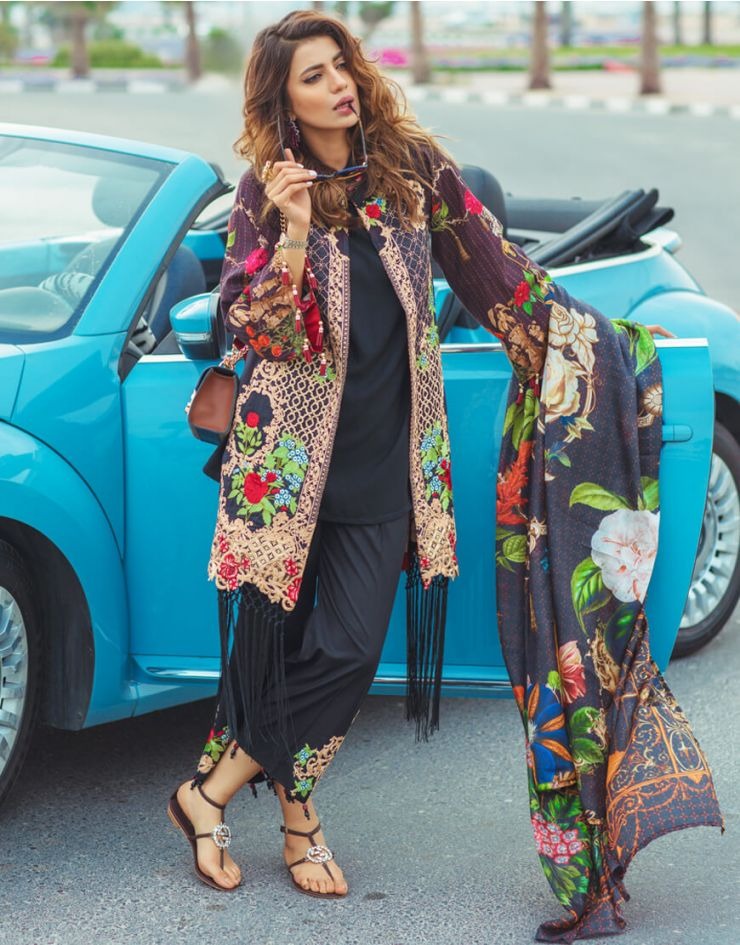 A woman posing in front of a bright blue convertible car, wearing a black pants set with a colorful floral and paisley print open-front jacket and a matching long scarf.