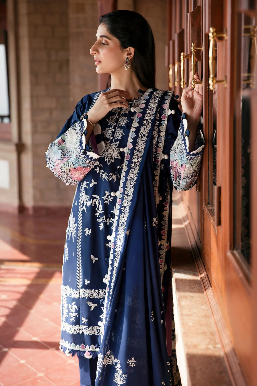 Woman in a blue embroidered traditional outfit standing in a sunlit room.