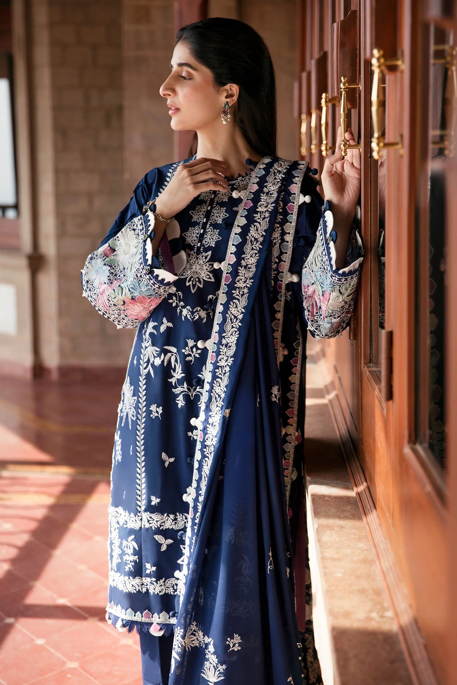 Woman in a blue embroidered traditional outfit standing in a sunlit room.