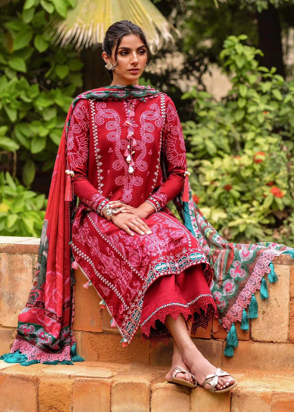 Woman in a red embroidered traditional outfit sitting outdoors with greenery in the background