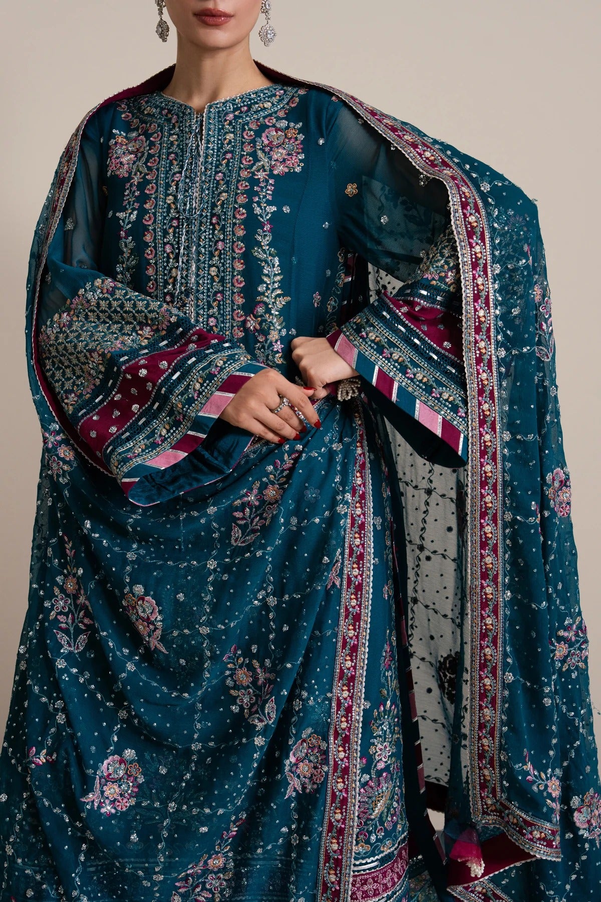 A close-up of a model wearing a traditional Indian salwar kameez suit in deep teal blue, featuring intricate silver and gold embroidery, floral prints, and a matching patterned dupatta.
