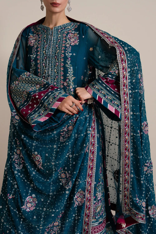 A close-up of a model wearing a traditional Indian salwar kameez suit in deep teal blue, featuring intricate silver and gold embroidery, floral prints, and a matching patterned dupatta.