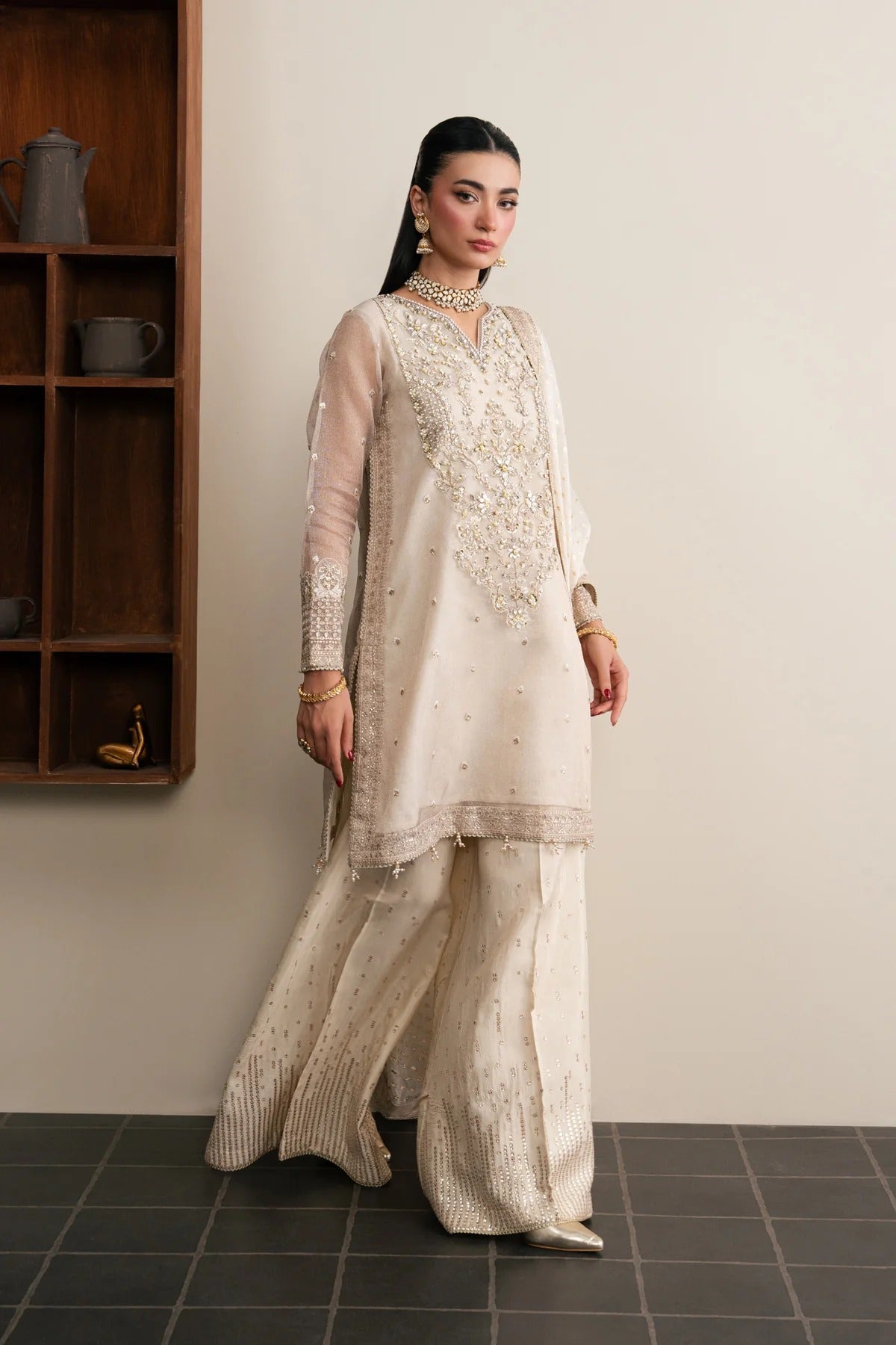 A woman models a traditional South Asian outfit consisting of a cream-colored embroidered kurta and matching wide-leg trousers, standing in an indoor setting with a wooden shelf on the left.
