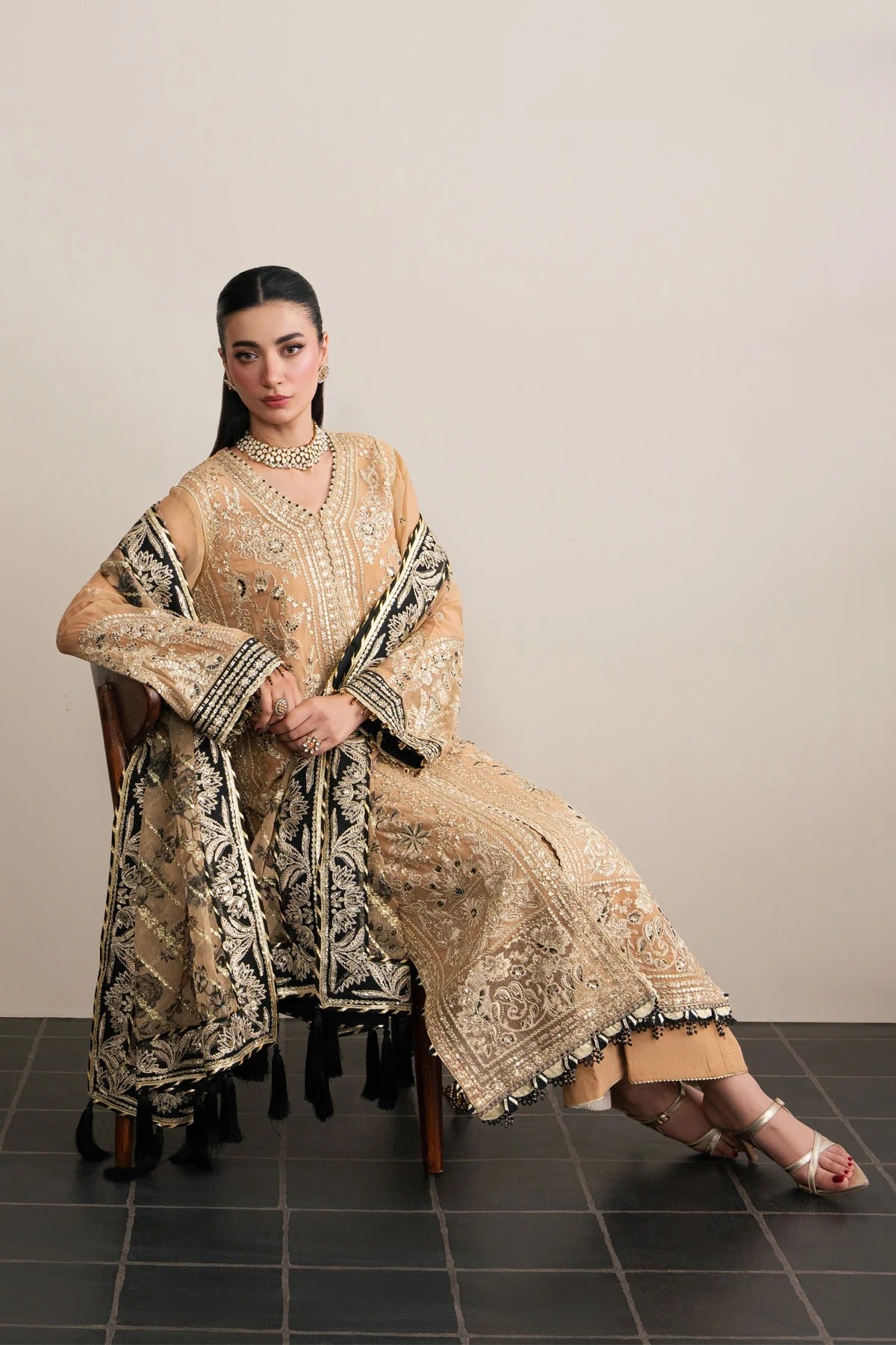 A woman seated on a wooden chair wearing an ornate beige and black embroidered traditional outfit with a matching draped scarf, photographed against a plain light beige wall and dark tiled floor.