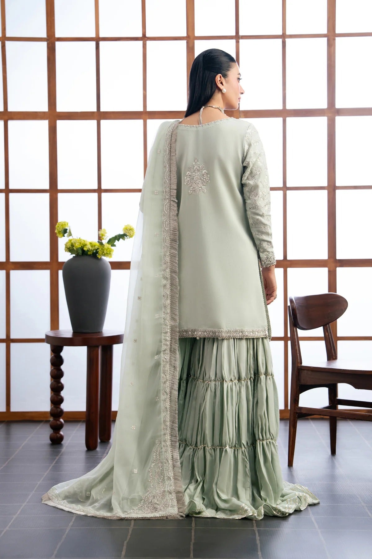 A woman stands with her back to the camera wearing a light sage green traditional outfit with a matching sheer dupatta, featuring intricate embroidery on the back and border details. She is posed in front of a wooden grid screen with a side table holding a vase of yellow flowers and a wooden chair.