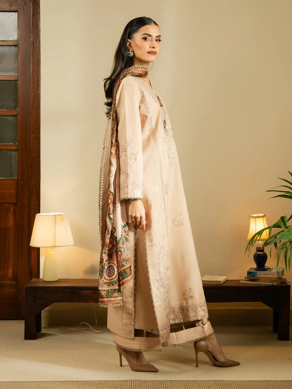 A woman models a traditional three-piece beige ethnic suit with matching printed dupatta in a warm-toned indoor setting with lit lamps and a wooden bench.