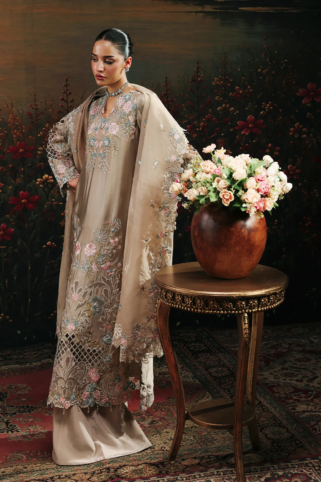 A woman models an elegant beige embroidered traditional outfit with a matching sheer dupatta, standing beside a small wooden table holding a large vase of pink and white flowers against a dark floral backdrop.