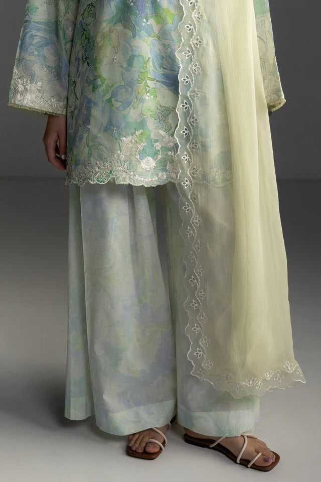 A model wearing a traditional South Asian outfit featuring a light blue and green floral printed kurta with white lace borders, matching wide-leg trousers, and a sheer cream dupatta, paired with brown sandals.